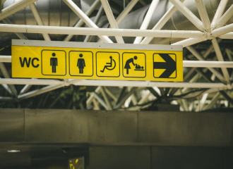 An overhead wayfinding sign in yellow in a transport building with pictograms for toilets, accessible toilet and baby change facilities